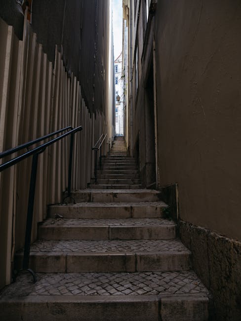 A person wearing a dark grey t-shirt and beige trousers is lifting a large, rectangular cardboard box with packing tape along the edges, inside a home with wooden stairs and a wooden handrail. The box