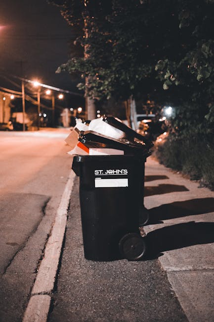 Three large plastic waste disposal containers are positioned outdoors in front of a modern residential building with yellow and white exterior walls. Two of the containers are yellow and one is green,